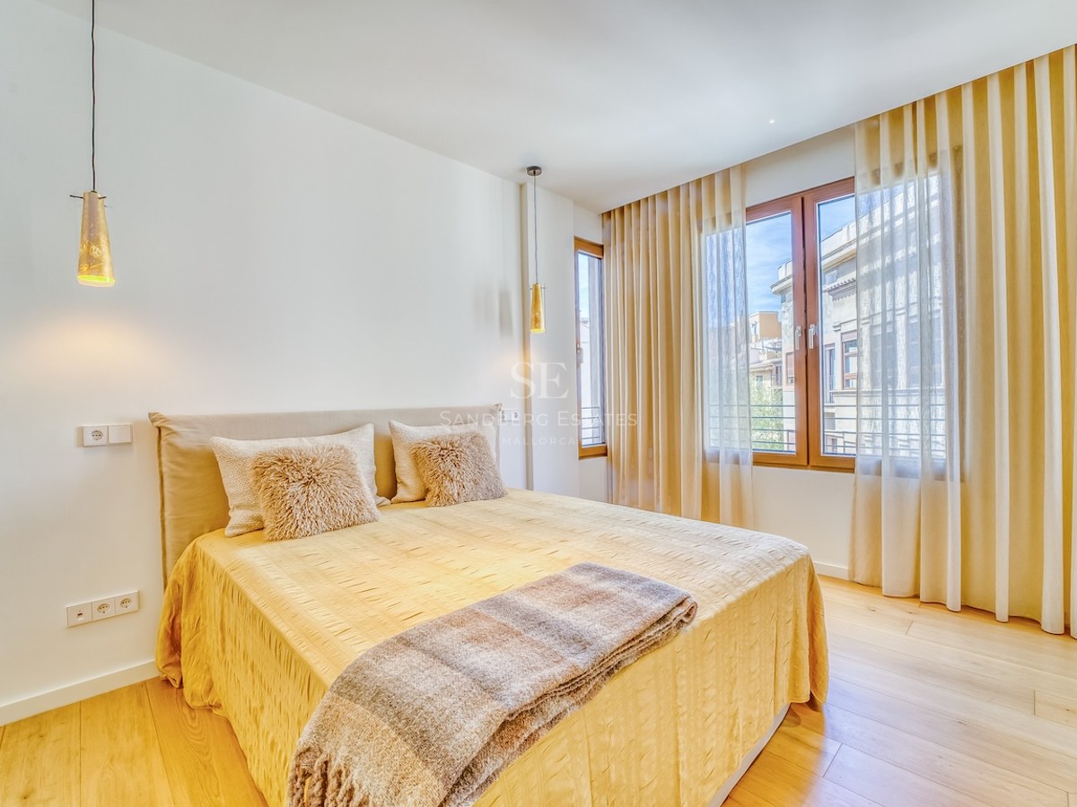 Bedroom with double bed, light wood floors, large windows, and beige curtains in a bright setting.