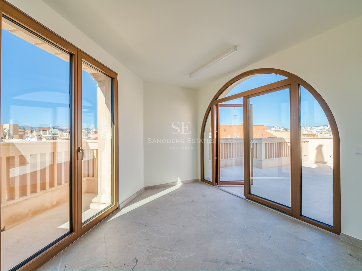 Empty room with marble floors and a large arched glass door opening to a sunny urban terrace under a clear blue sky.
