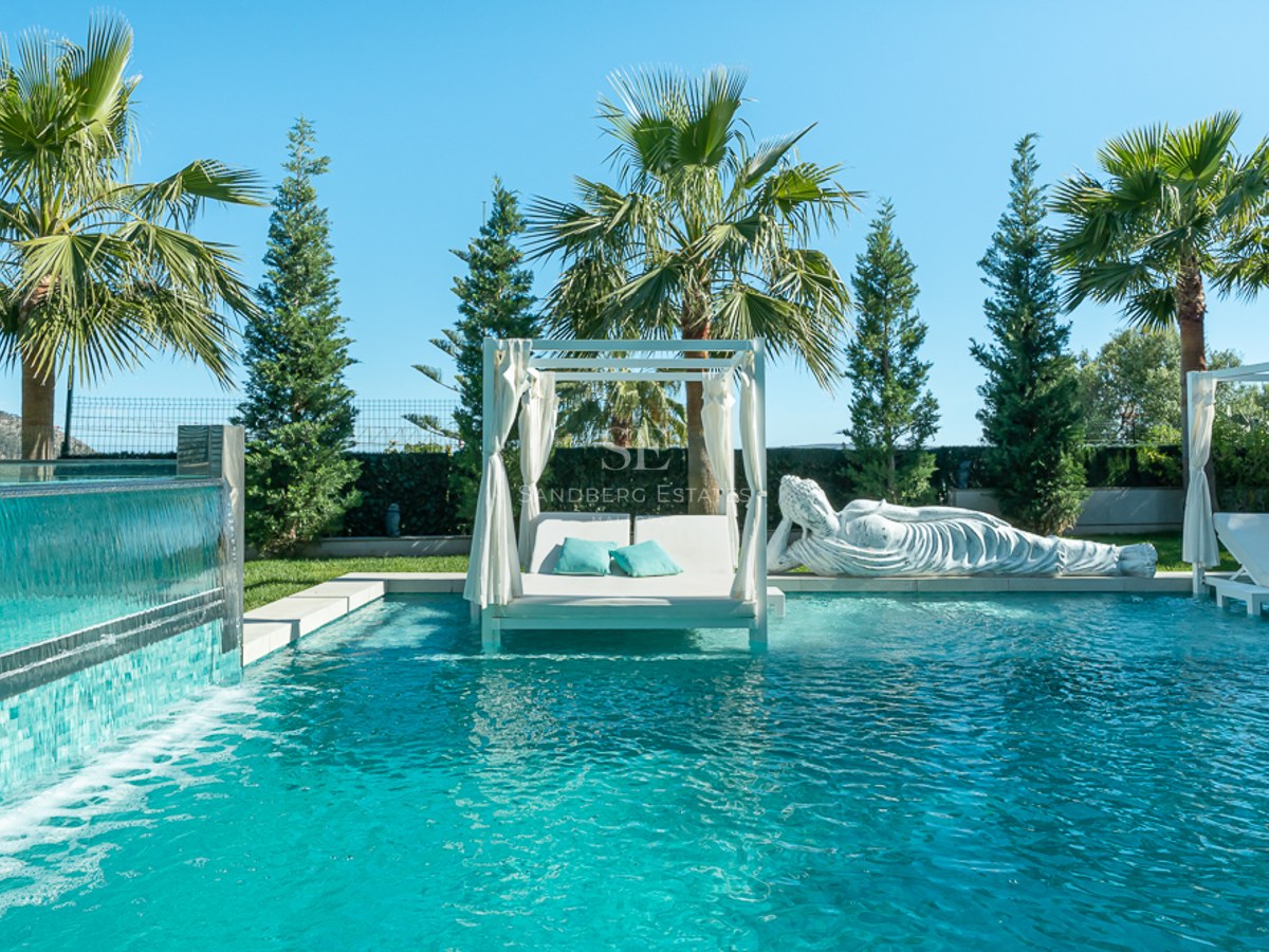 Vue sur piscine à débordement avec chaises longues, entourée d'un jardin luxuriant. Architecture moderne avec larges fenêtres et balcon.