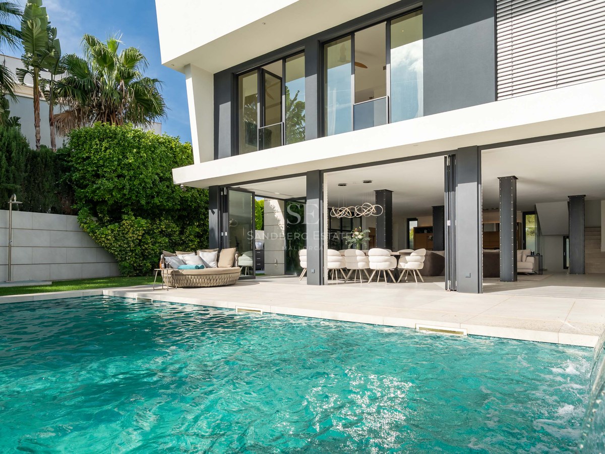 Extérieur de villa moderne avec piscine turquoise, cascade et salon ouvert sous un ciel bleu éclatant.