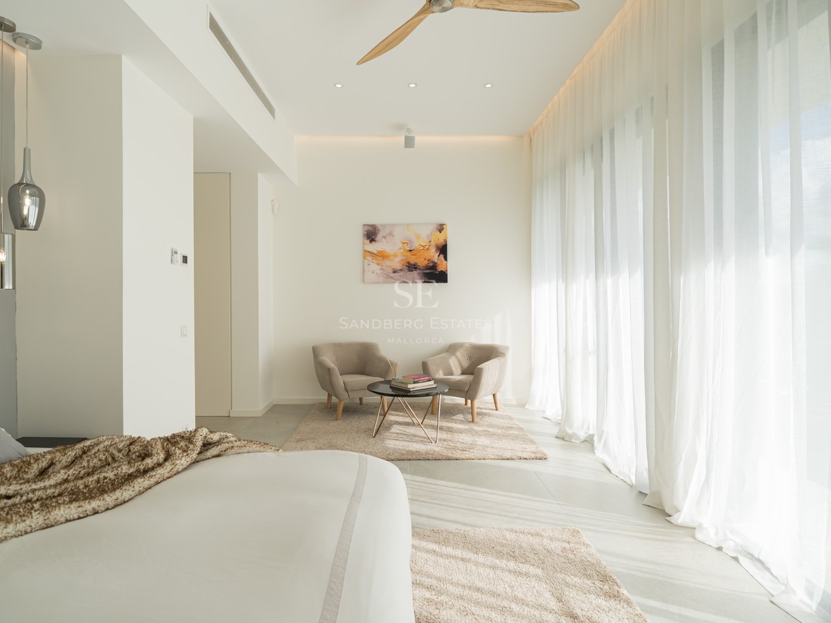 Grande chambre blanche avec baies vitrées, rideaux voilages, deux fauteuils et ventilateur de plafond en bois.