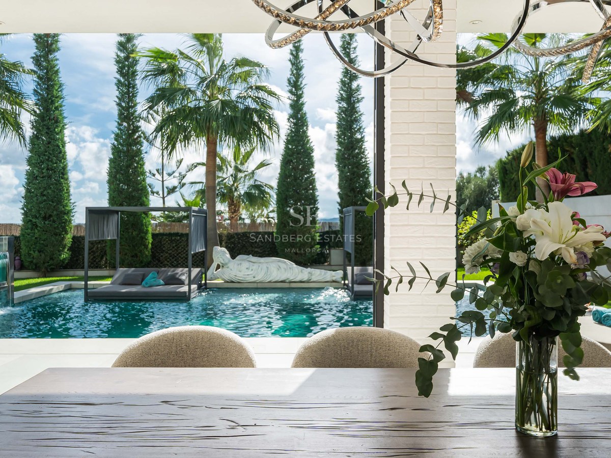 Table à manger en bois donnant sur une piscine turquoise et un jardin tropical avec palmiers et lits de jour.