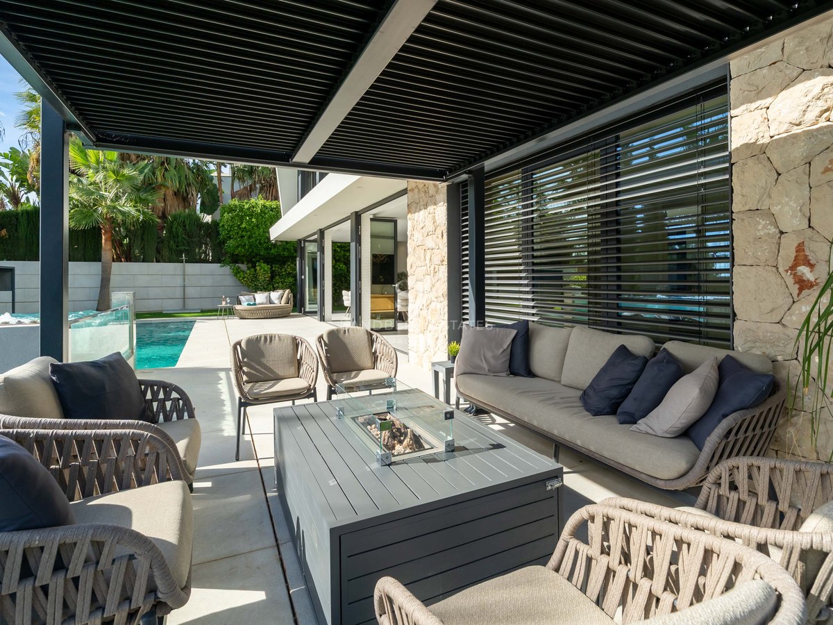 Salon extérieur de luxe avec mobilier gris sous une pergola noire, à côté d'une piscine et d'un mur en pierre.