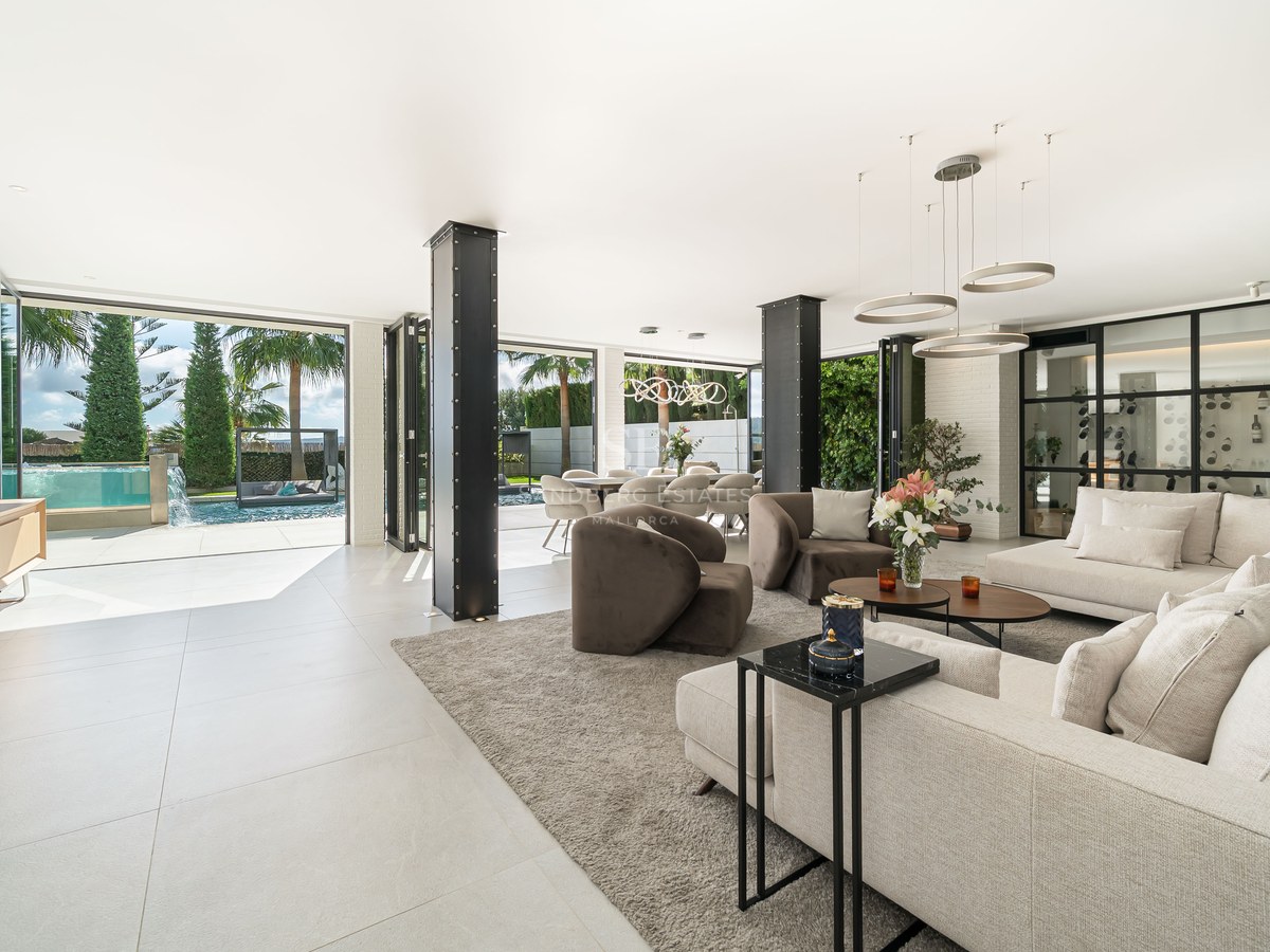 Salon moderne spacieux avec piliers en acier, canapés blancs et grandes baies vitrées ouvrant sur une piscine.
