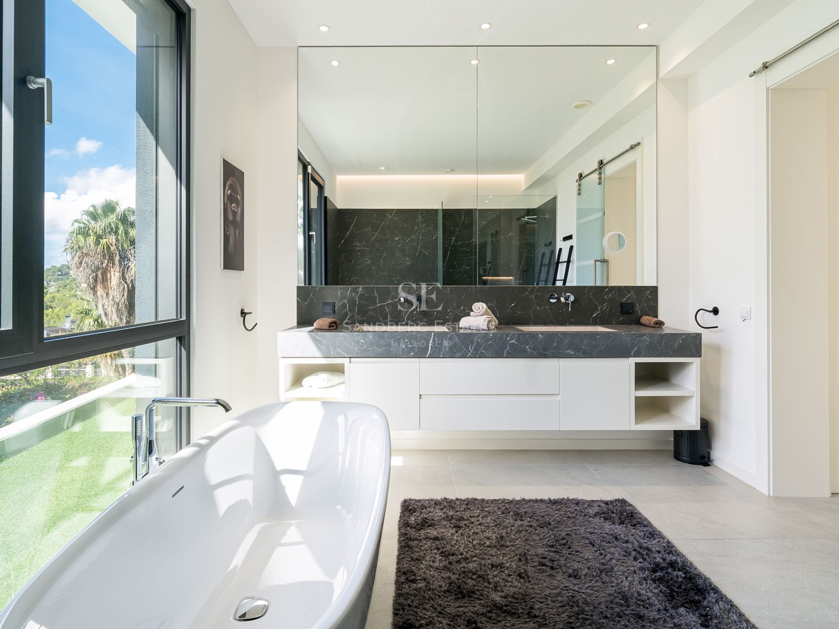Salle de bain contemporaine lumineuse avec baignoire îlot près d'une grande fenêtre donnant sur un jardin verdoyant.