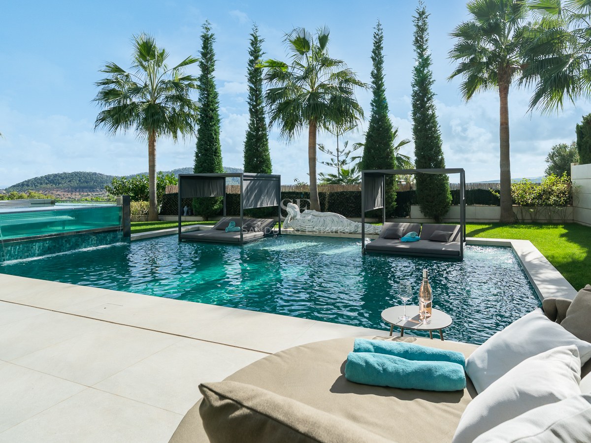 Piscine de luxe avec paroi en verre, lits intégrés, palmiers et vue sur la montagne sous un ciel bleu.