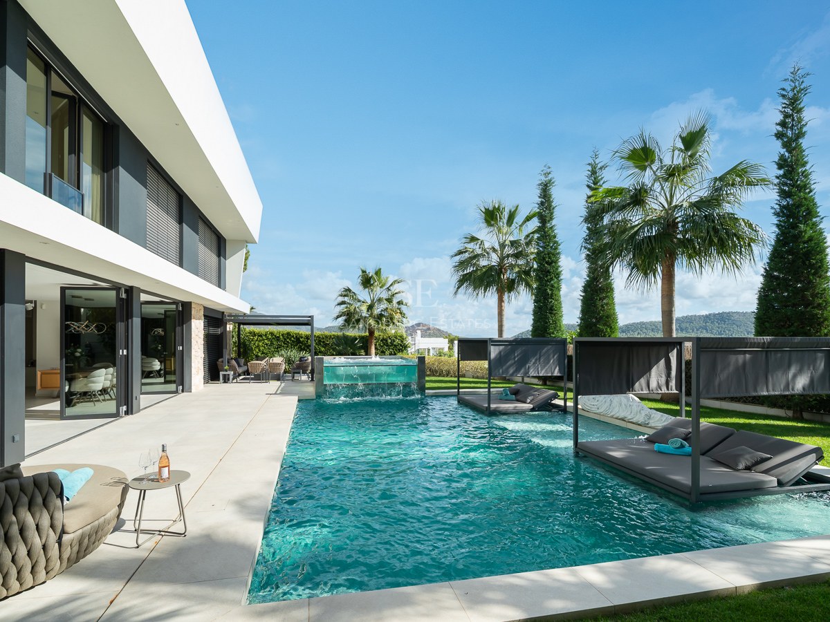 Grande piscine turquoise avec lits de repos immergés et cascade en verre devant une villa blanche moderne.