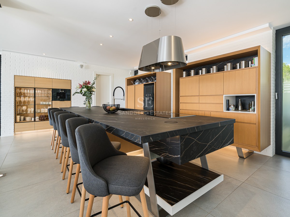 Cuisine moderne avec un grand îlot en marbre noir, tabourets gris, meubles en bois clair et mur en briques blanches.
