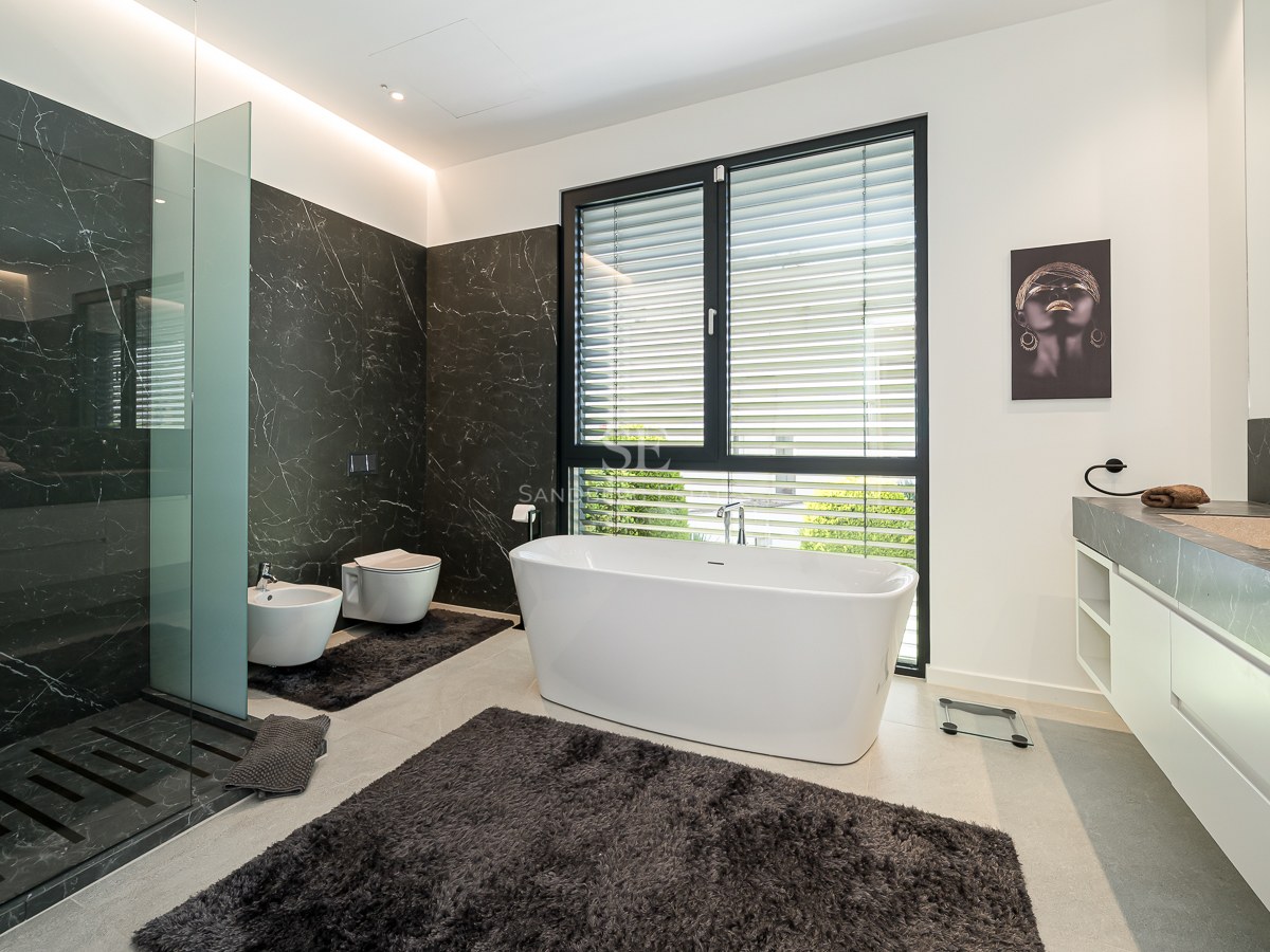 Salle de bains moderne avec baignoire blanche îlot, douche en marbre sombre et grande fenêtre avec stores.