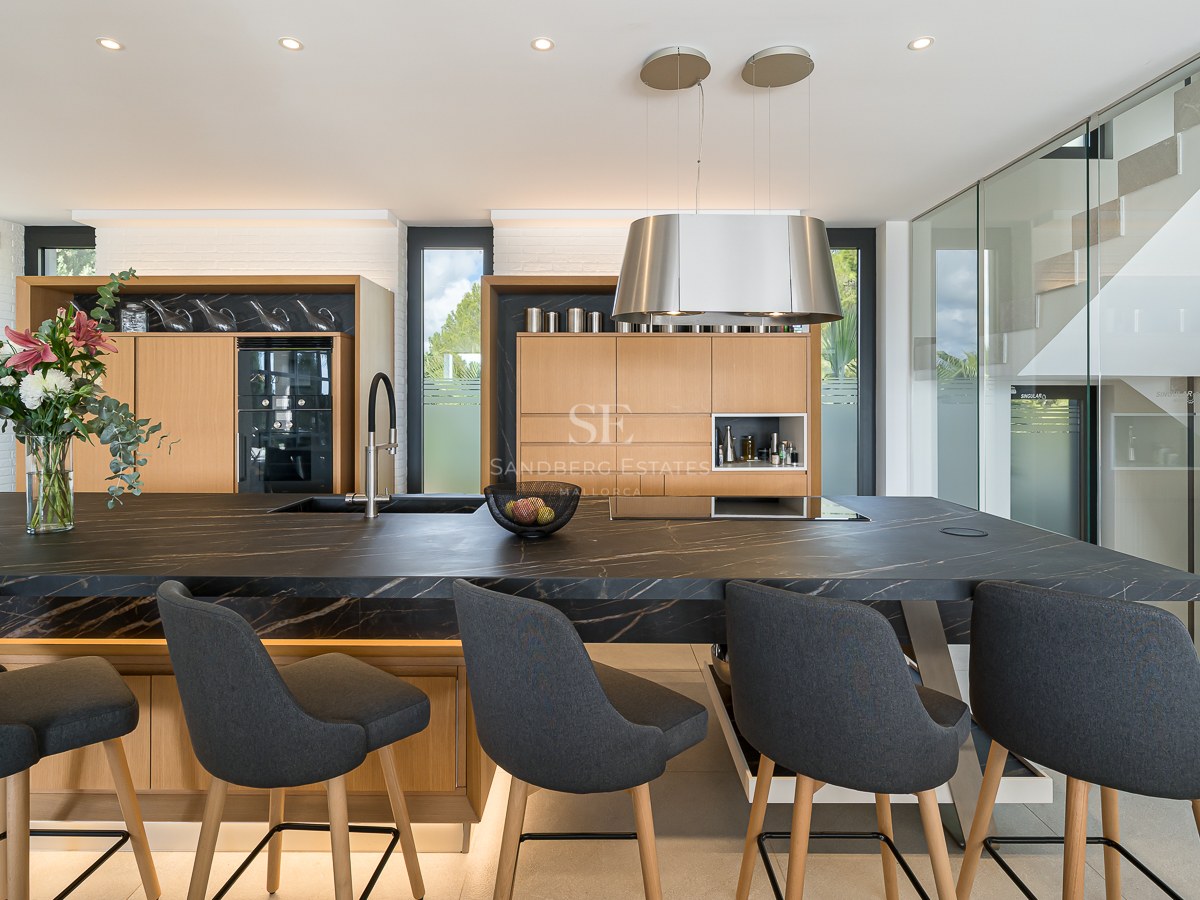 Cuisine moderne avec un grand îlot en pierre noire, tabourets gris, meubles en chêne et escalier en verre.