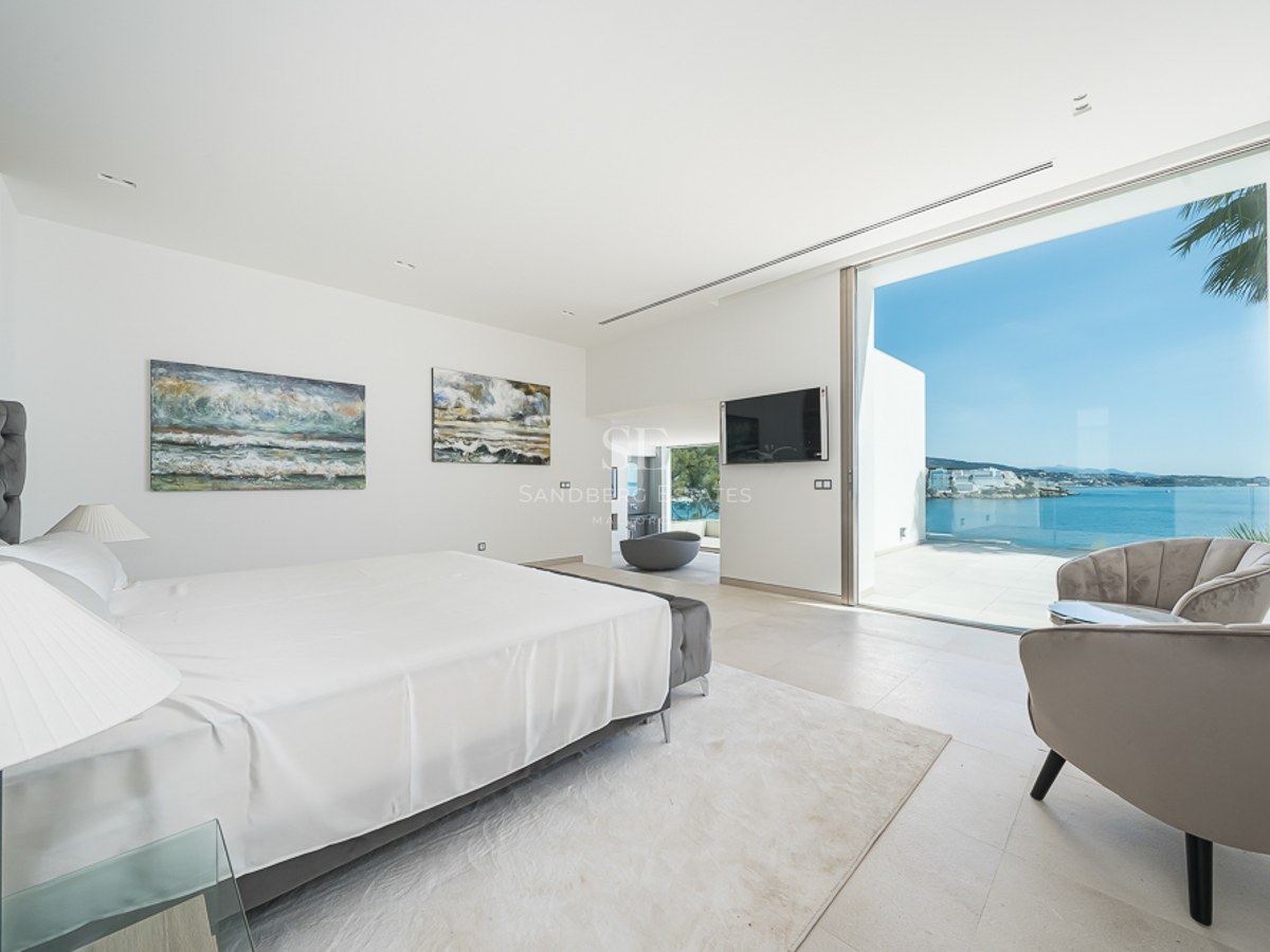 Dormitorio luminoso con cama blanca, sillones de terciopelo y grandes ventanales con vistas al mar Mediterráneo.
