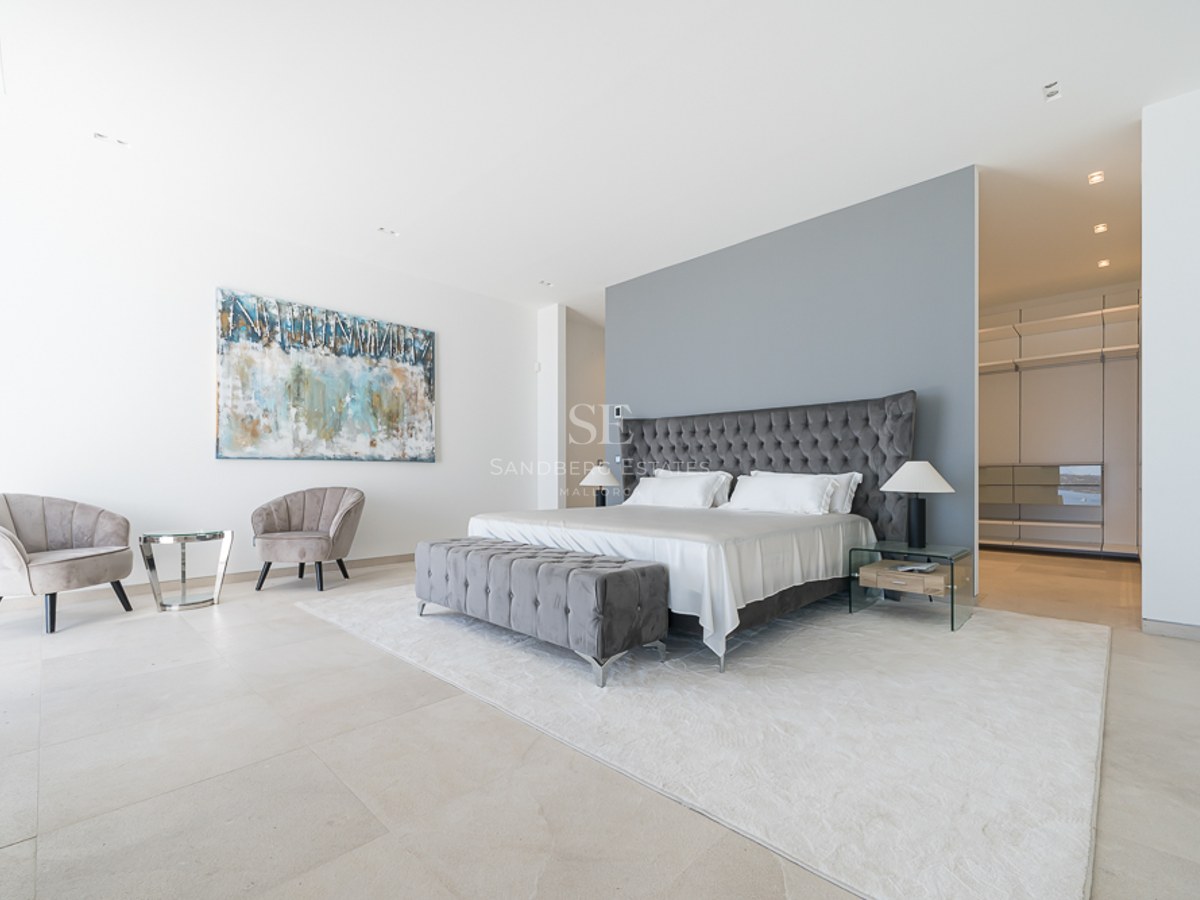 Dormitorio principal con cama capitoné gris, zona de estar de terciopelo y vestidor al fondo.
