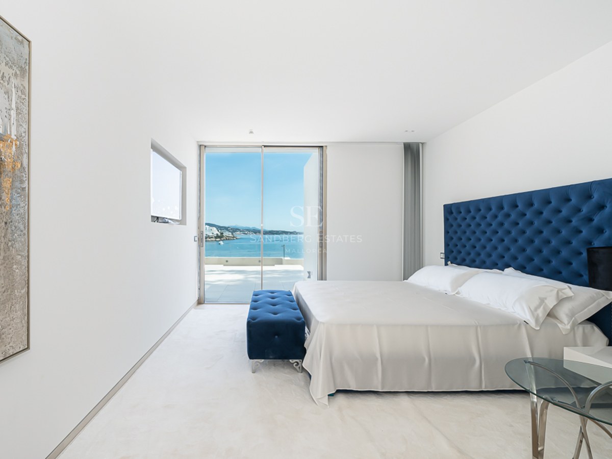 Dormitorio moderno con cabecero capitoné azul, alfombra blanca y grandes ventanales con vistas al océano.