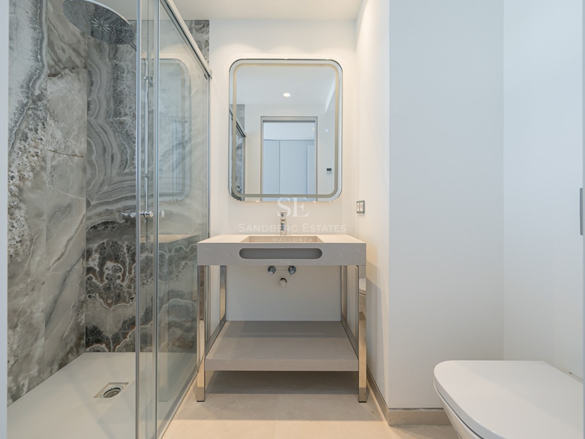 Baño moderno con ducha de cristal y paredes de mármol, espejo retroiluminado y lavabo minimalista.
