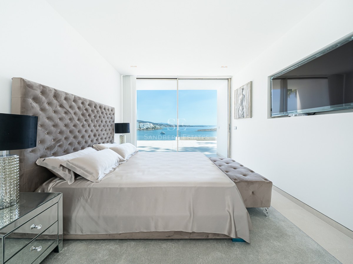 Dormitorio moderno con cabecero capitoné, muebles de espejo y grandes ventanales con vistas al océano azul.