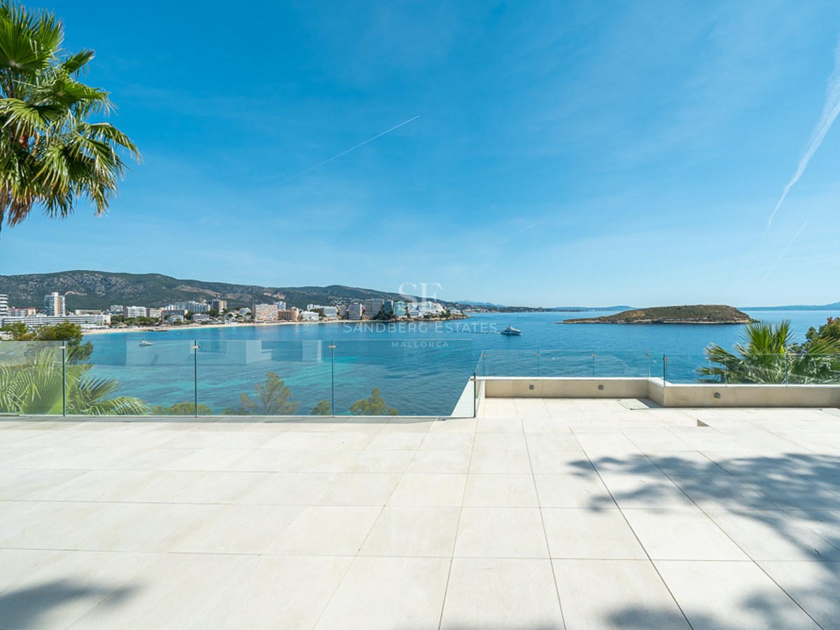 Amplia terraza de baldosas blancas con barandillas de cristal con vistas a una bahía azul y la costa.