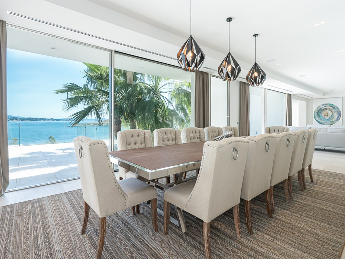 Elegante comedor con mesa larga, sillas tapizadas de color beige y grandes ventanales con vistas al mar y palmeras.