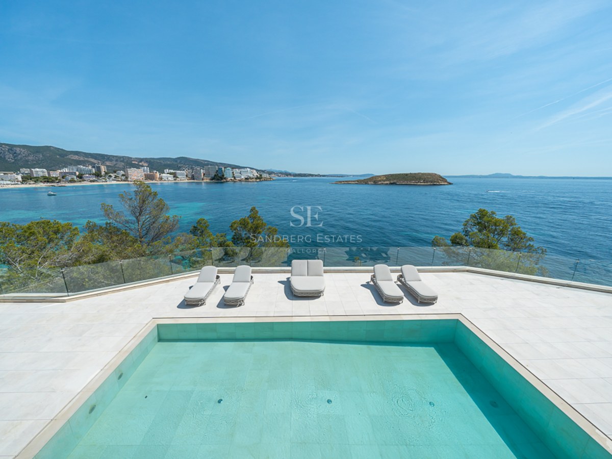 Piscina moderna y tumbonas en una terraza blanca con vistas a una bahía turquesa y la costa.