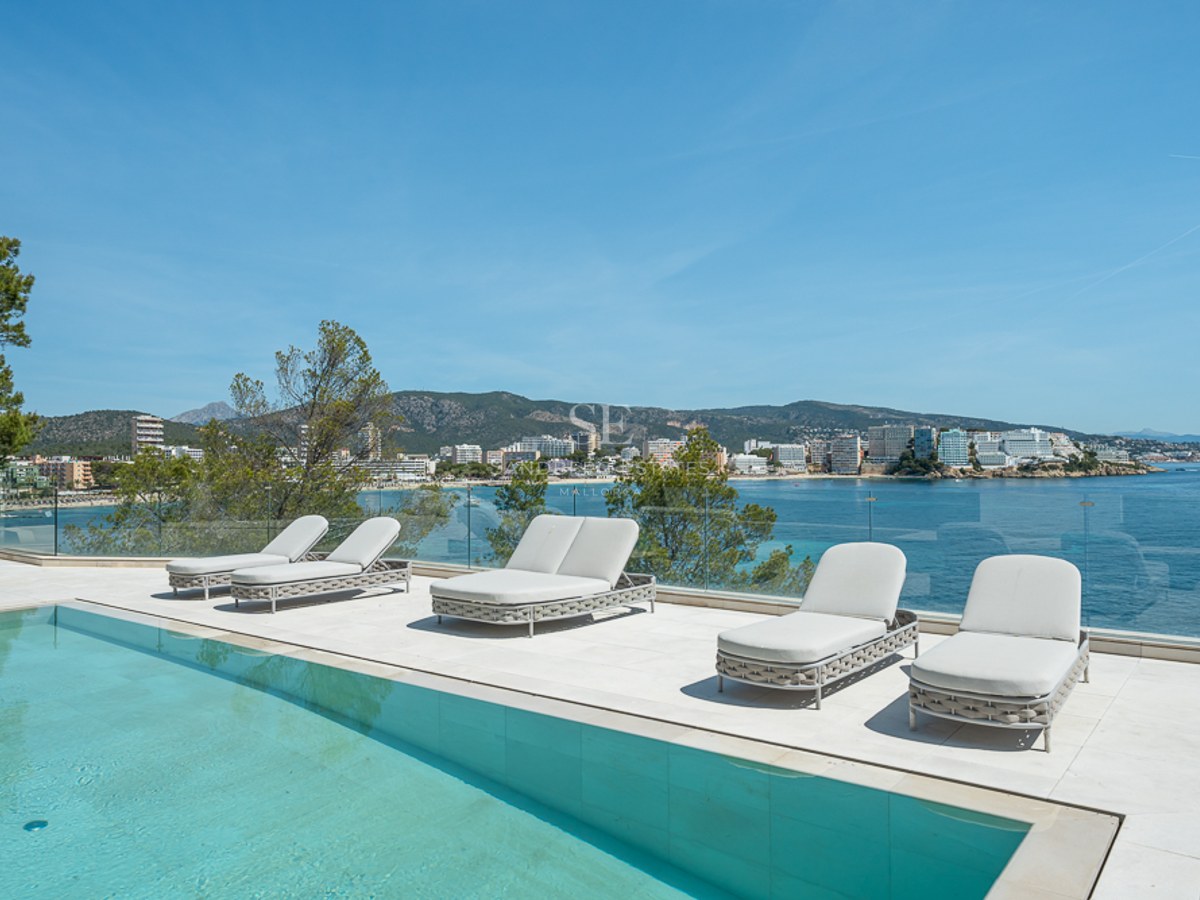 Terraza blanca moderna con piscina turquesa, tumbonas y barandilla de cristal con vistas a una bahía mediterránea.