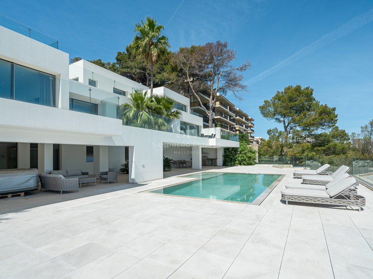 Villa blanca moderna con una piscina rectangular, tumbonas grises y barandillas de cristal bajo un cielo azul.