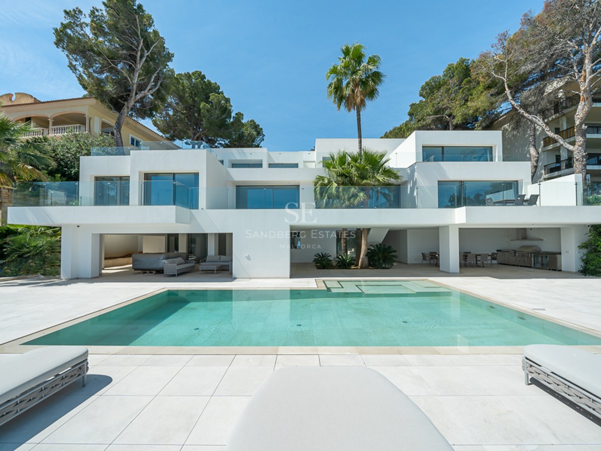 Villa moderna blanca con una gran piscina turquesa, barandillas de cristal y palmeras bajo un cielo despejado.