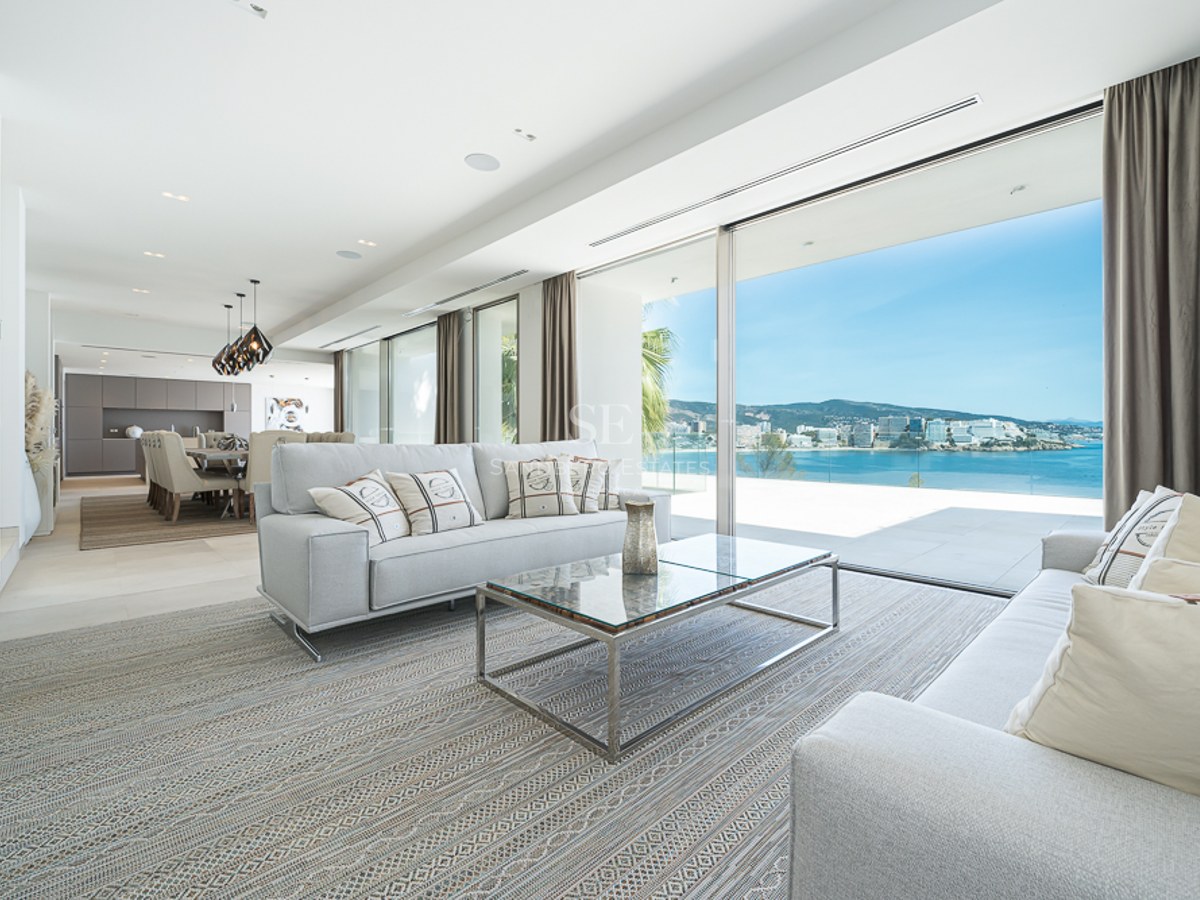 Salón moderno con sofás grises y grandes ventanales que muestran una vista panorámica del mar y la costa.