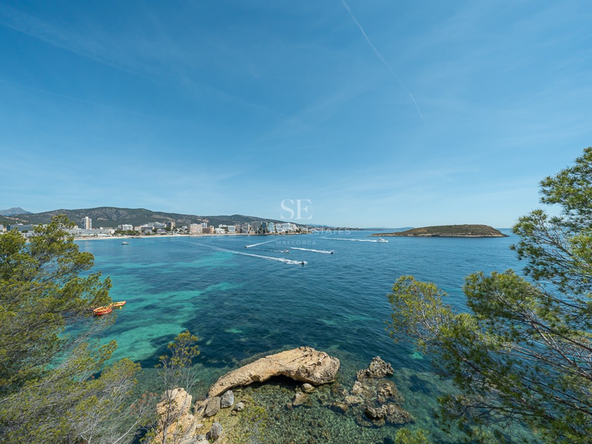 Vista de una bahía de aguas turquesas con barcos, rodeada de pinos y una ciudad costera al fondo.