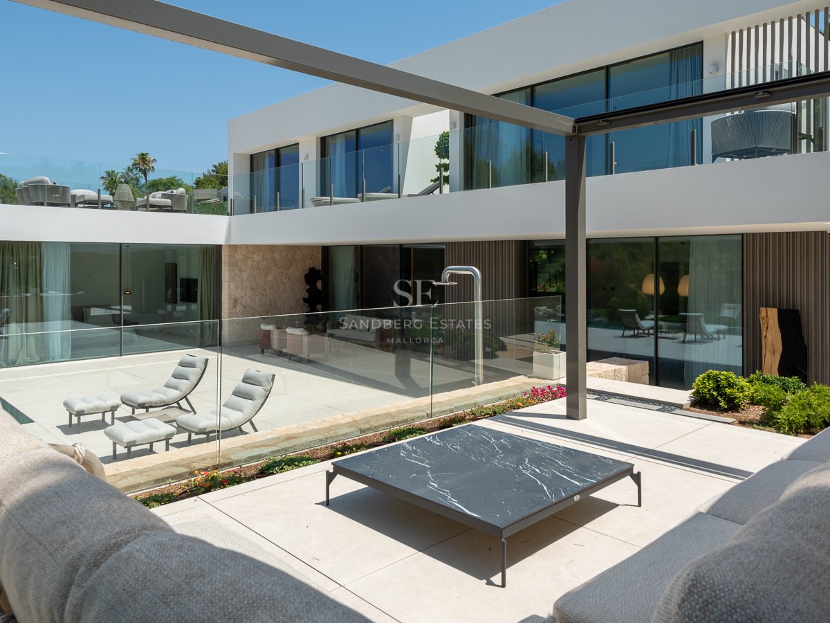 Villa blanche moderne avec balustrades en verre, murs en pierre et terrasse ensoleillée avec mobilier de salon design.