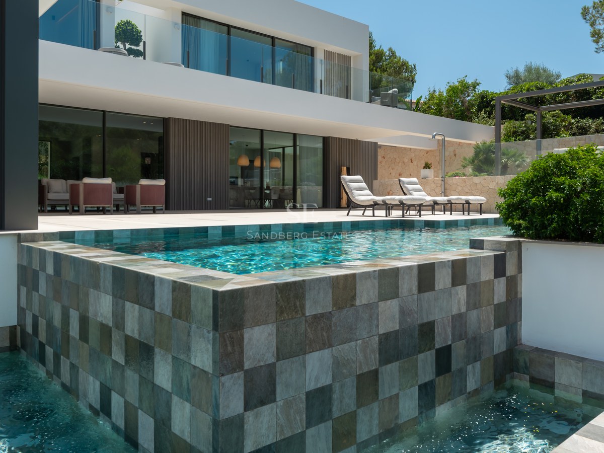 Villa blanche moderne avec piscine à débordement en pierre, chaises longues et grandes baies vitrées.