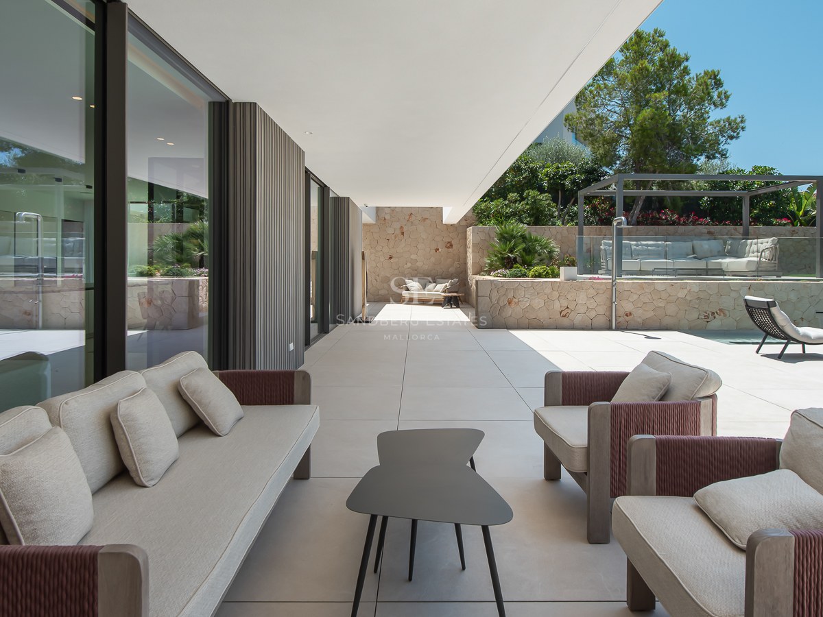 Terrasse couverte avec mobilier gris, murs en pierre naturelle et piscine sous un ciel bleu.