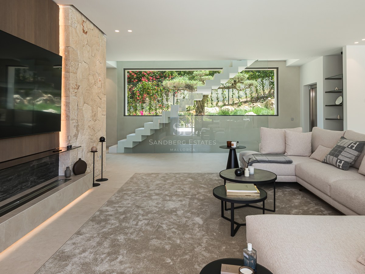 Salon moderne avec canapé beige, mur en pierre avec cheminée intégrée et escalier en verre vue sur jardin.