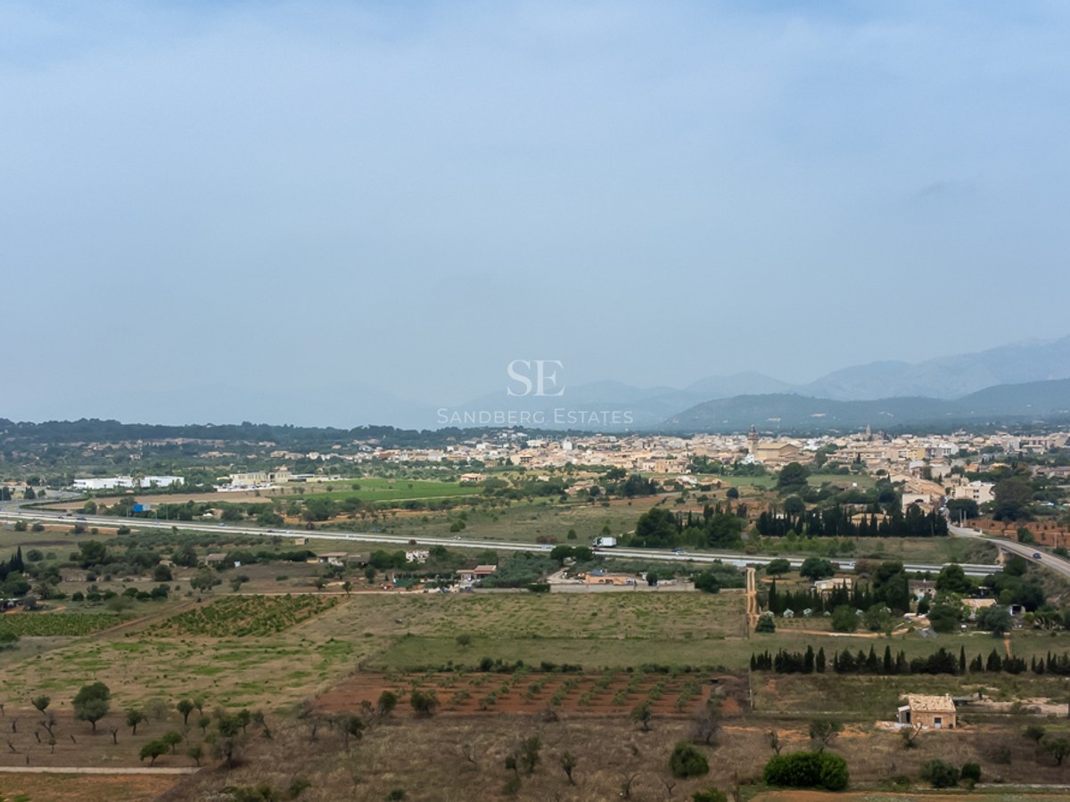 Weite Landschaft einer mallorquinischen Stadt, umgeben von Feldern und Bergen unter dunstigem blauem Himmel.