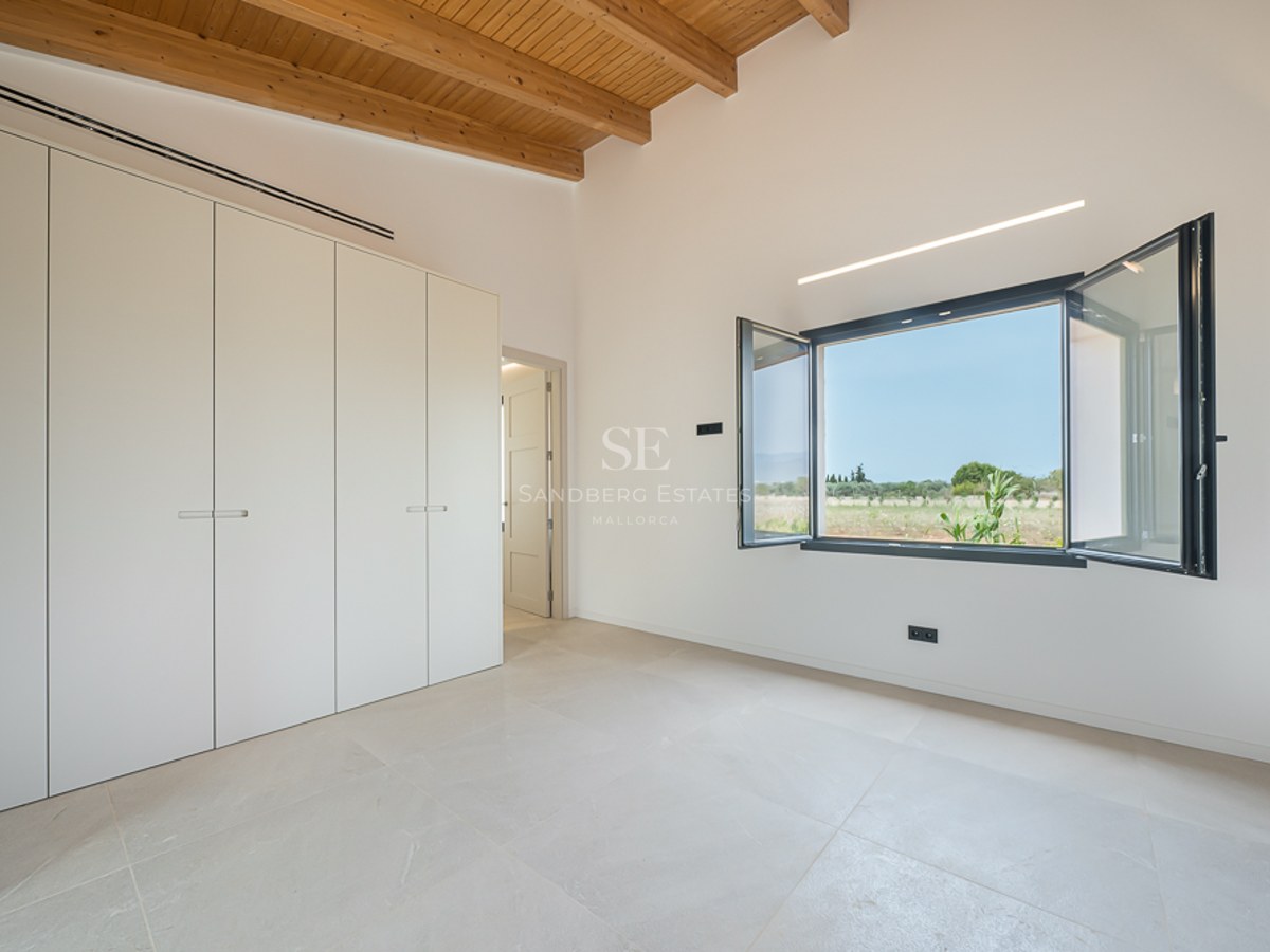 Helles Schlafzimmer mit hohen Holzbalkendecken, weißen Einbauschränken und offenem Fenster mit Naturblick.