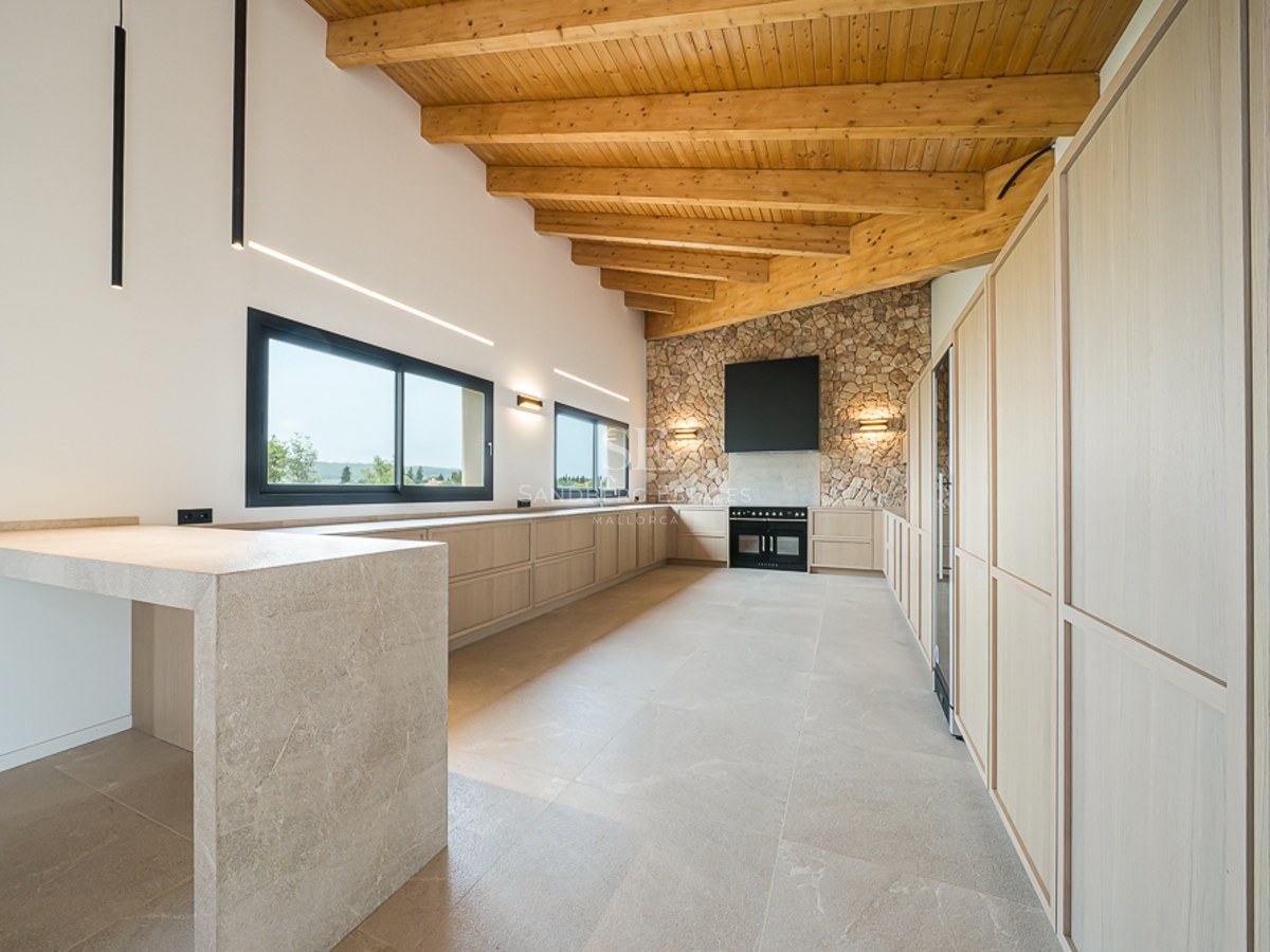 Geräumige moderne Küche mit hohen Holzbalkendecken, Natursteinwand, beigen Schränken und schwarzen Geräten.