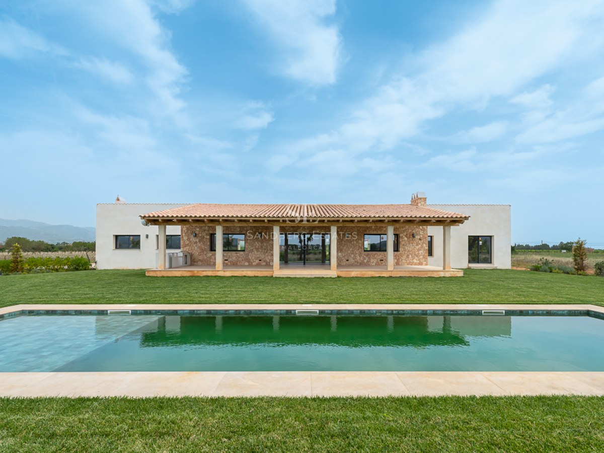 Luxusvilla mit langem rechteckigem Pool, grünem Rasen, Steinfassade und Terrakotta-Veranda unter blauem Himmel.