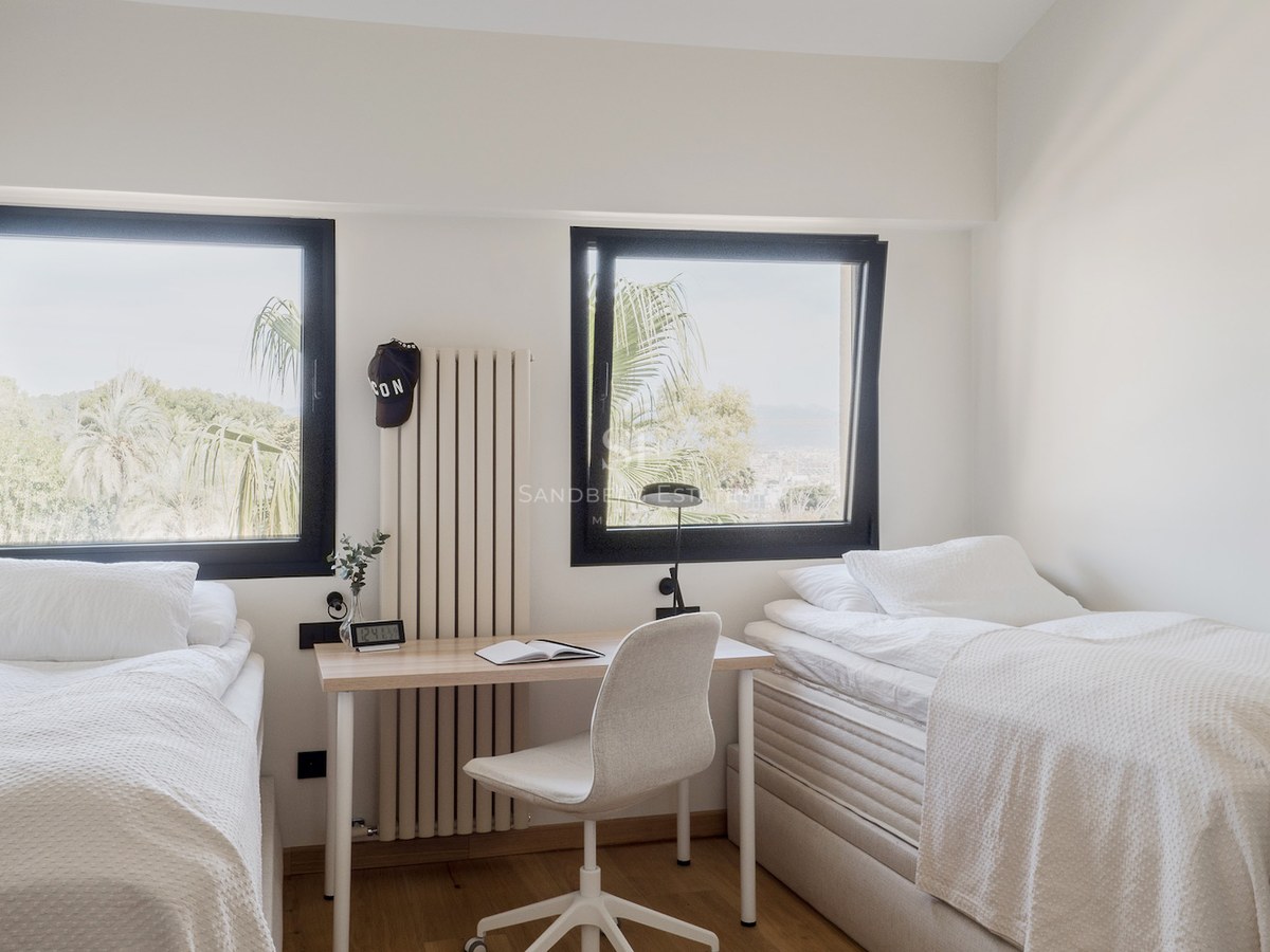 Dormitorio con dos camas individuales blancas, un escritorio central y dos ventanas con vistas a palmeras.