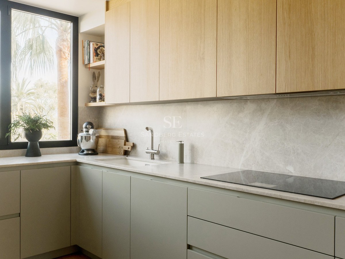 Cocina de diseño con armarios de madera clara, encimera de piedra gris y ventana con vistas a palmeras.
