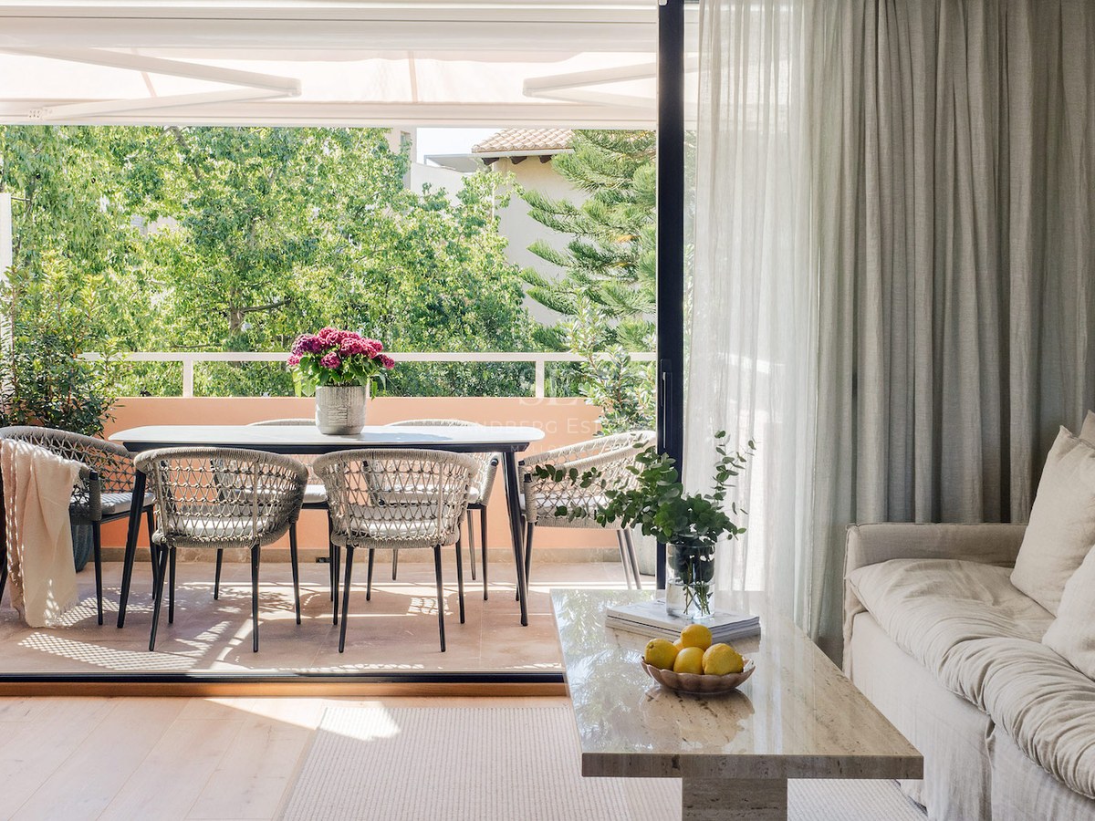 Vista desde un salón luminoso hacia una terraza amueblada con mesa de comedor, rodeada de frondosos árboles verdes.