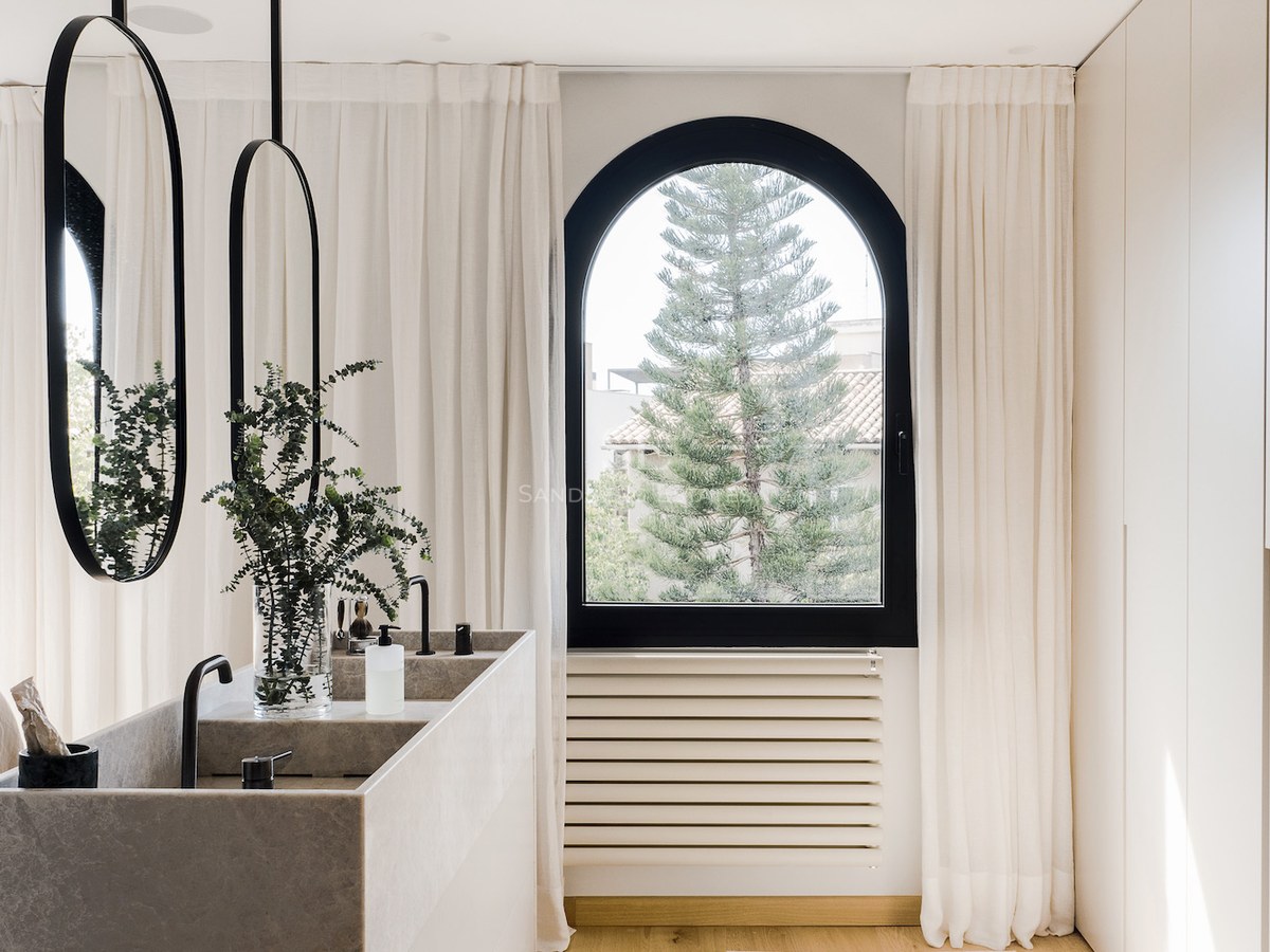 Lavabo doble de piedra con espejos ovalados negros frente a una ventana de arco con vistas a un árbol.
