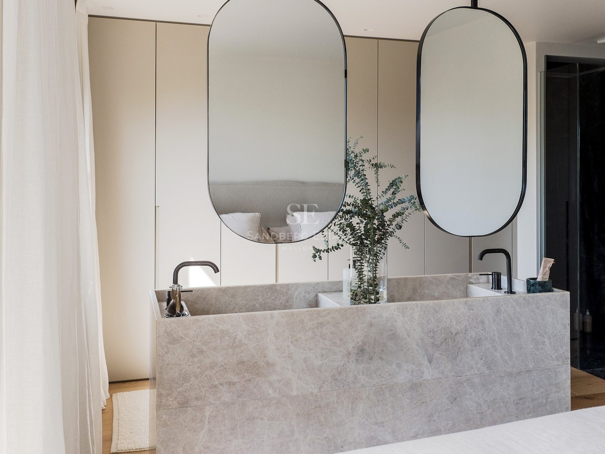 Lavabo doble moderno de piedra mármol, dos espejos ovalados y grifos negros en un baño minimalista y luminoso.