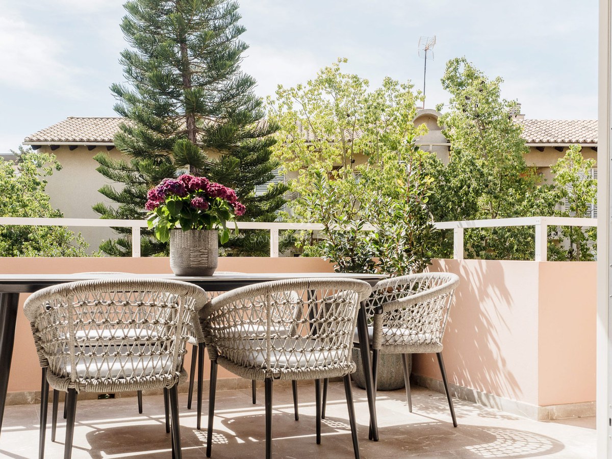 Terraza soleada con mesa negra, sillas tejidas y flores púrpuras, con vistas a árboles exuberantes.
