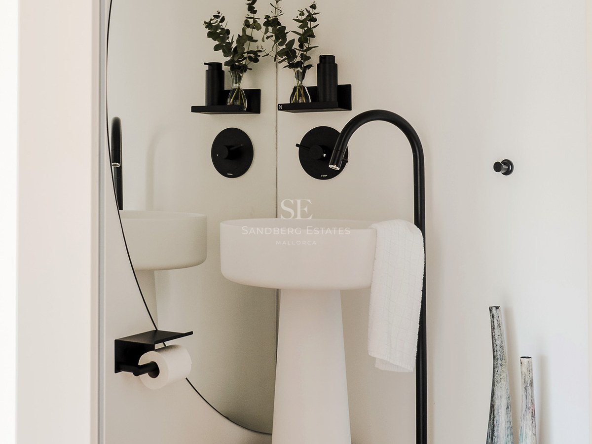 Lavabo de pedestal blanco con grifo negro mate alto, espejo redondo y ramas de eucalipto.