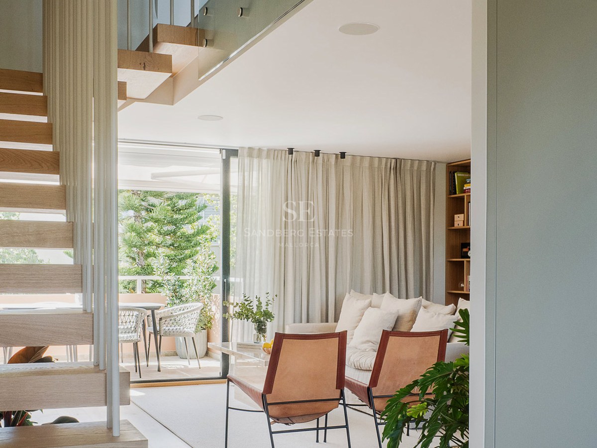 Vista de un salón luminoso con escaleras flotantes de madera, sillas de cuero y acceso a una terraza verde.