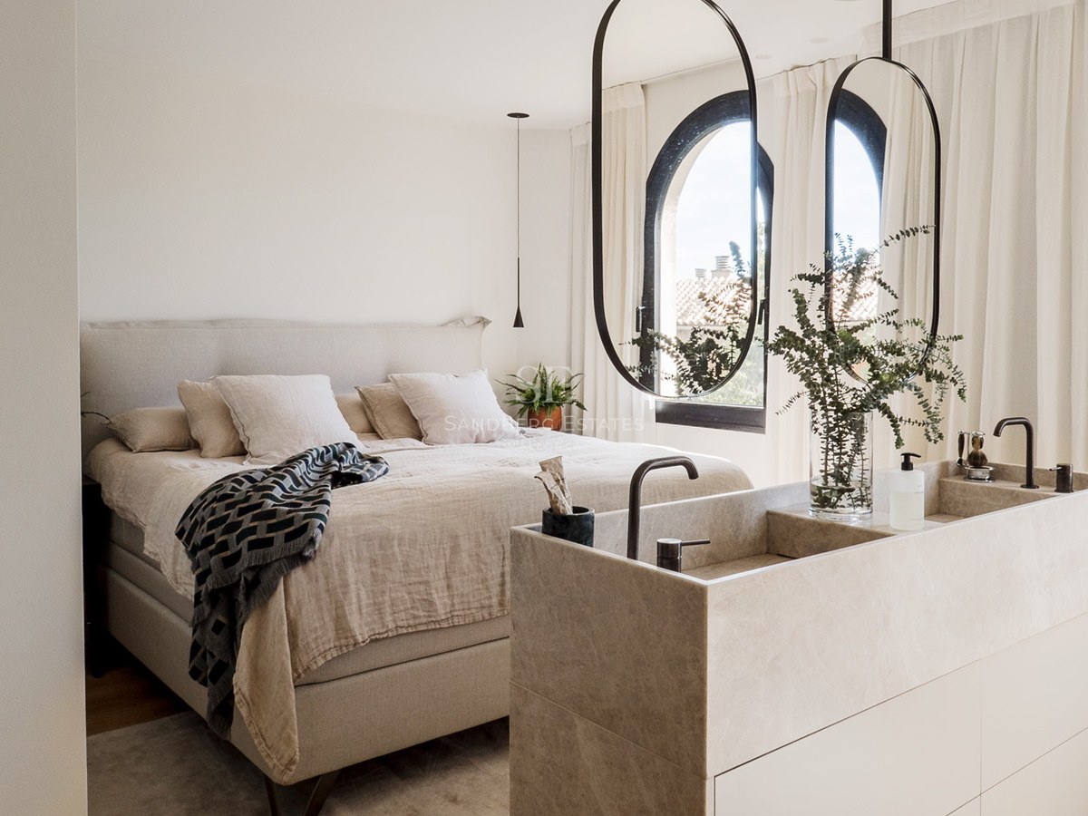 Dormitorio principal moderno con cama de tonos neutros y tocador de piedra integrado con espejos ovalados colgantes.