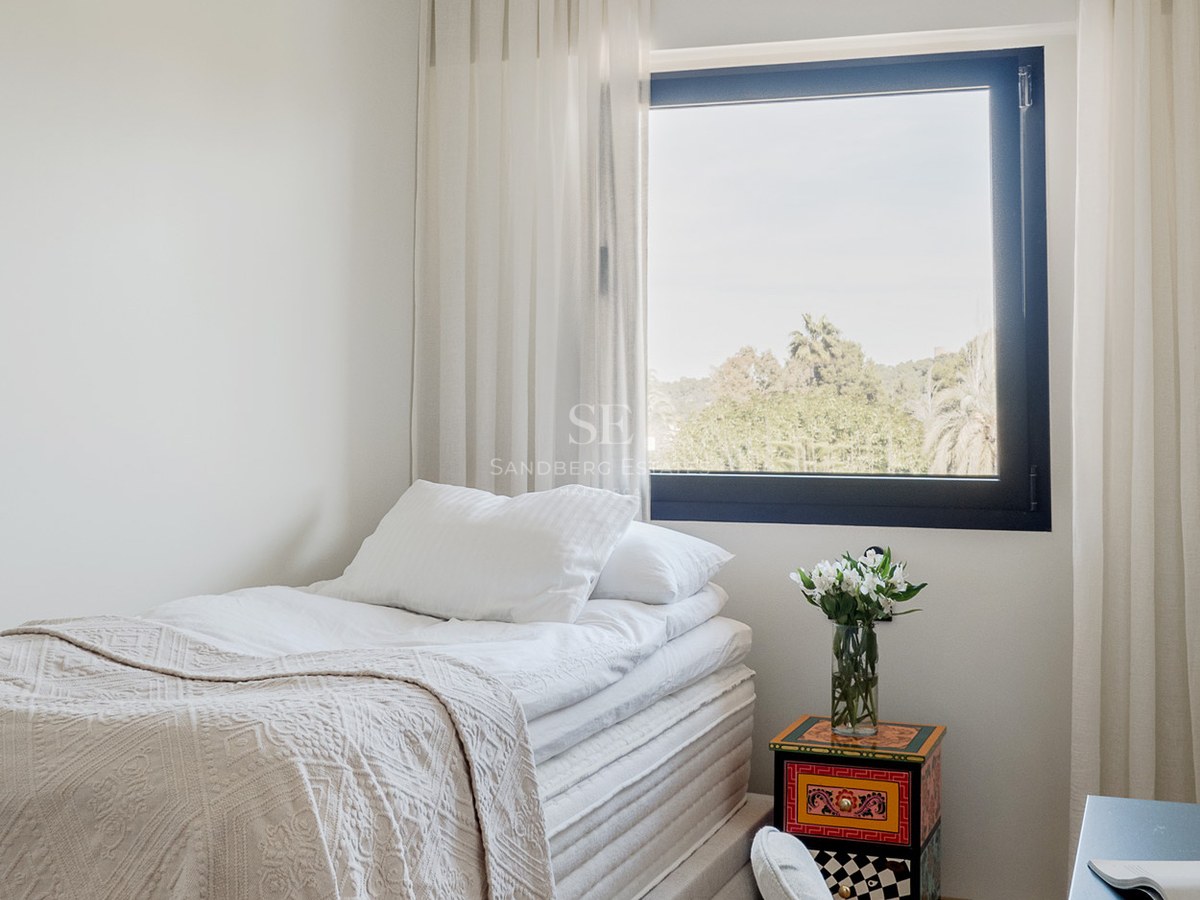 Cama individual con sábanas blancas junto a una ventana con vistas a árboles y una mesita de noche colorida.