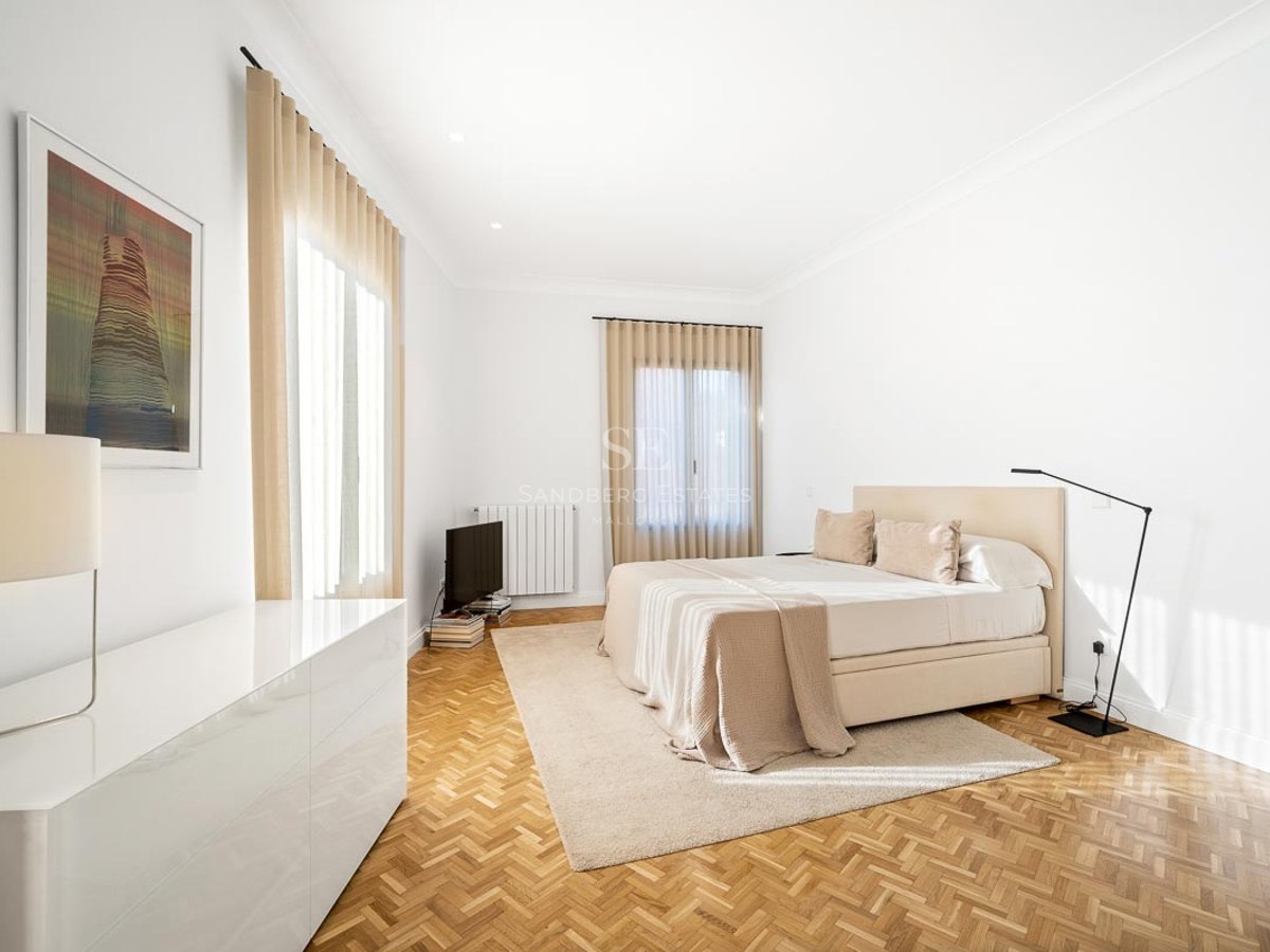 Chambre lumineuse avec parquet en chevrons, grand lit, rideaux beiges et lampe design.
