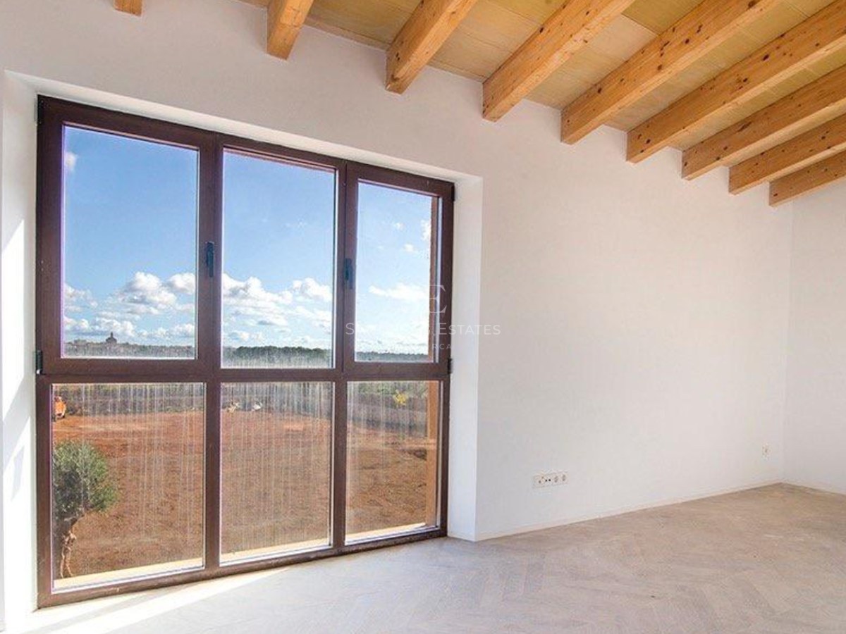 Empty room featuring exposed wooden ceiling beams and a large dark-framed window overlooking a sunny rural plot.