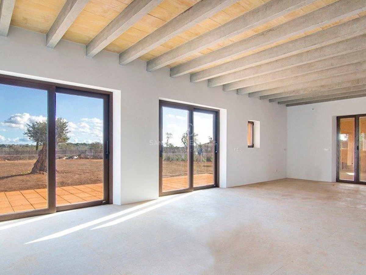 Spacious empty room with exposed concrete beam ceiling and sliding glass doors opening to a terrace with an olive tree.