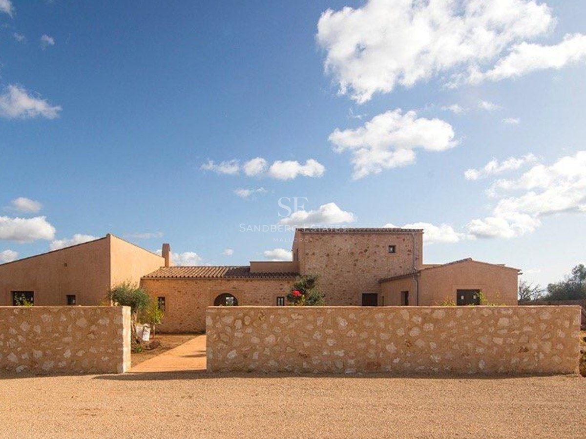 Exterior view of a stone and ochre finca under a blue sky, featuring a traditional stone wall and gravel entrance.