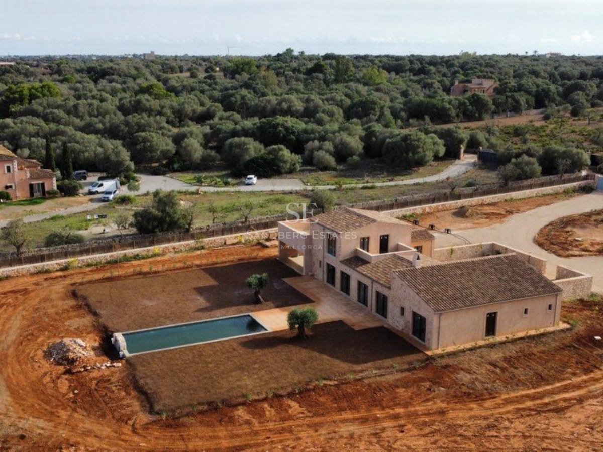 Aerial view of a newly built stone villa with a long swimming pool surrounded by a large plot and lush trees.